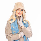 A woman wearing a beige knitted bucket hat with a blue scarf and a grey sweater, posing against a white background.