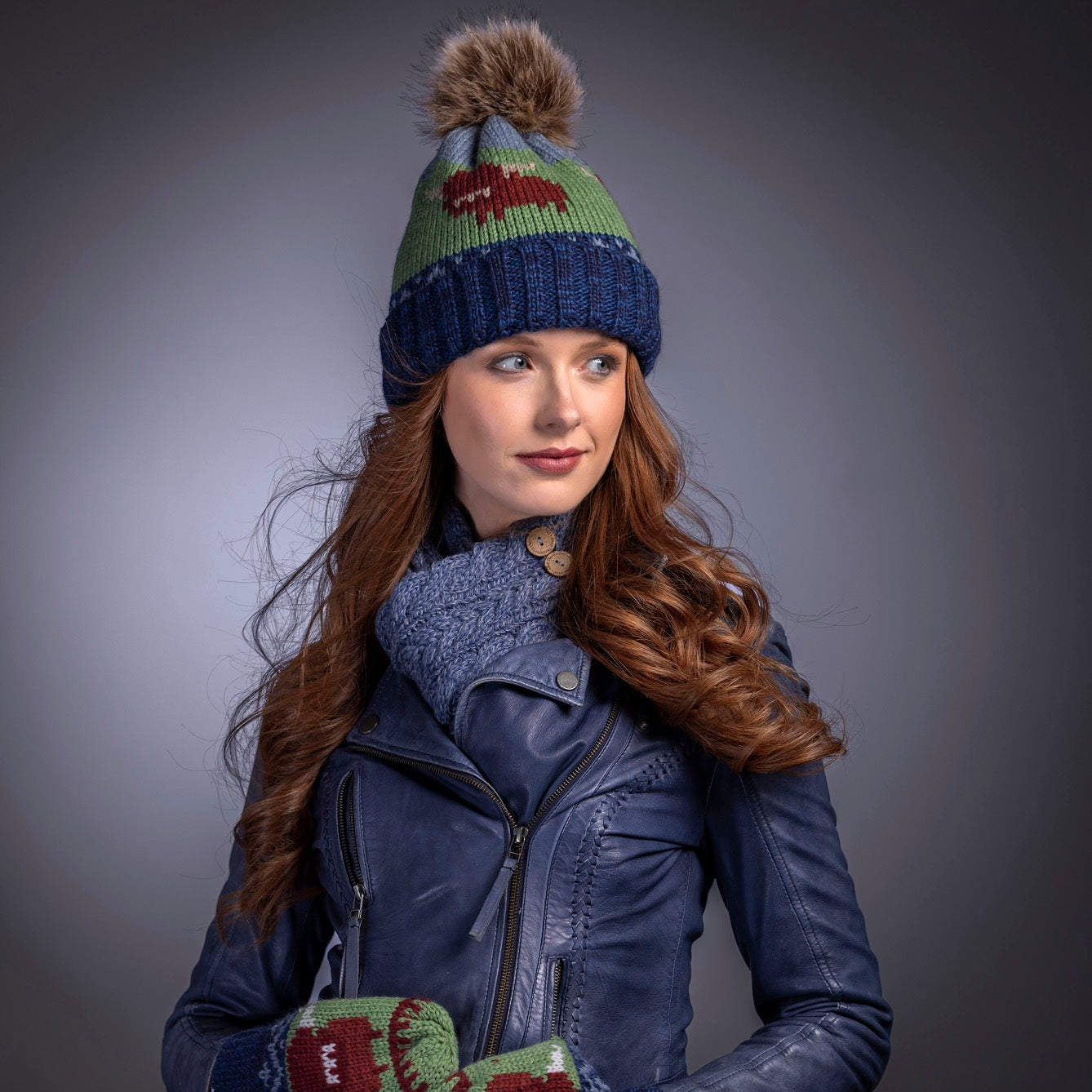 Woman wearing a blue knit hat with pom-pom, leather jacket, jeans, and colorful mittens against a gray background