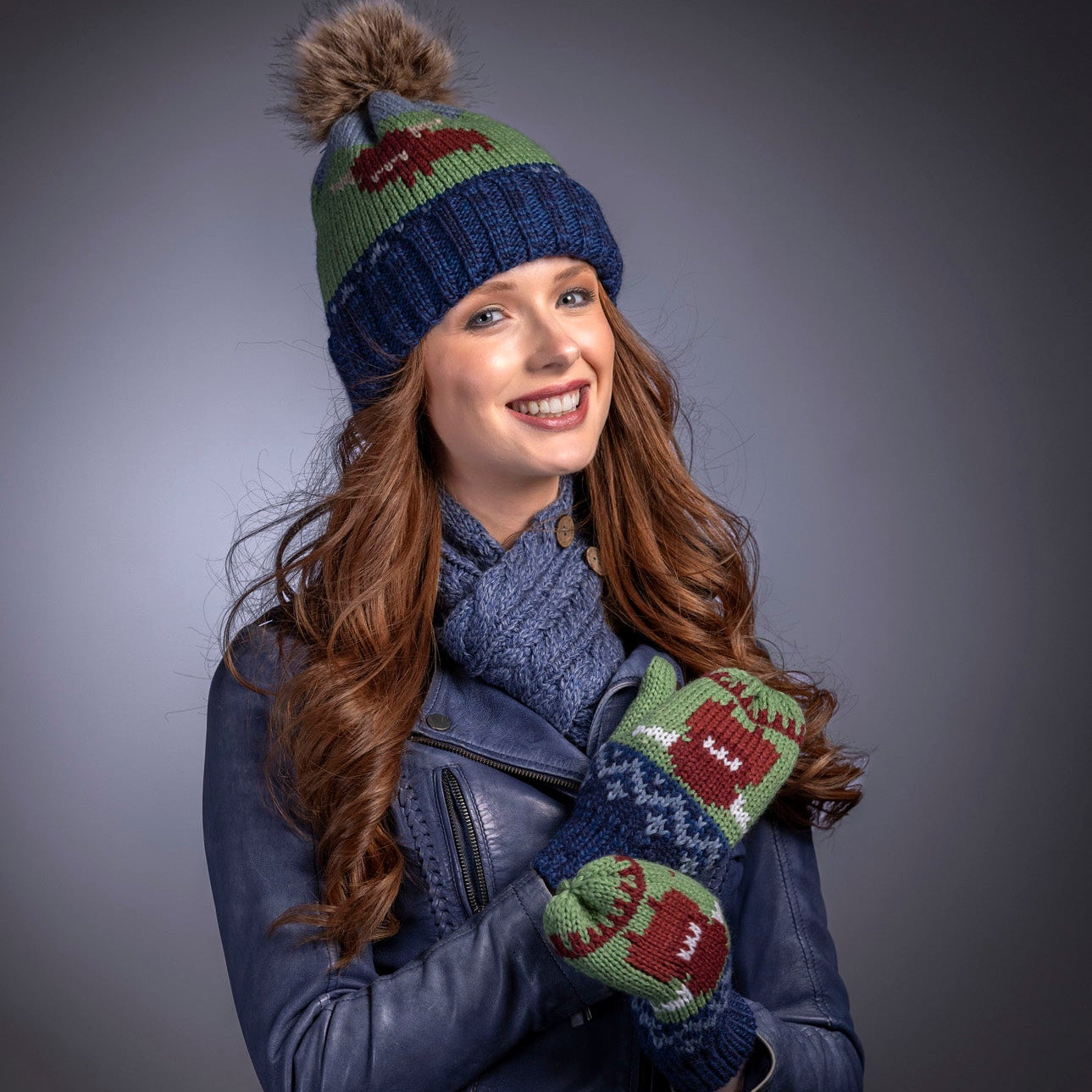 Woman wearing a blue knit hat, scarf, and mittens with highland cow design on a grey background