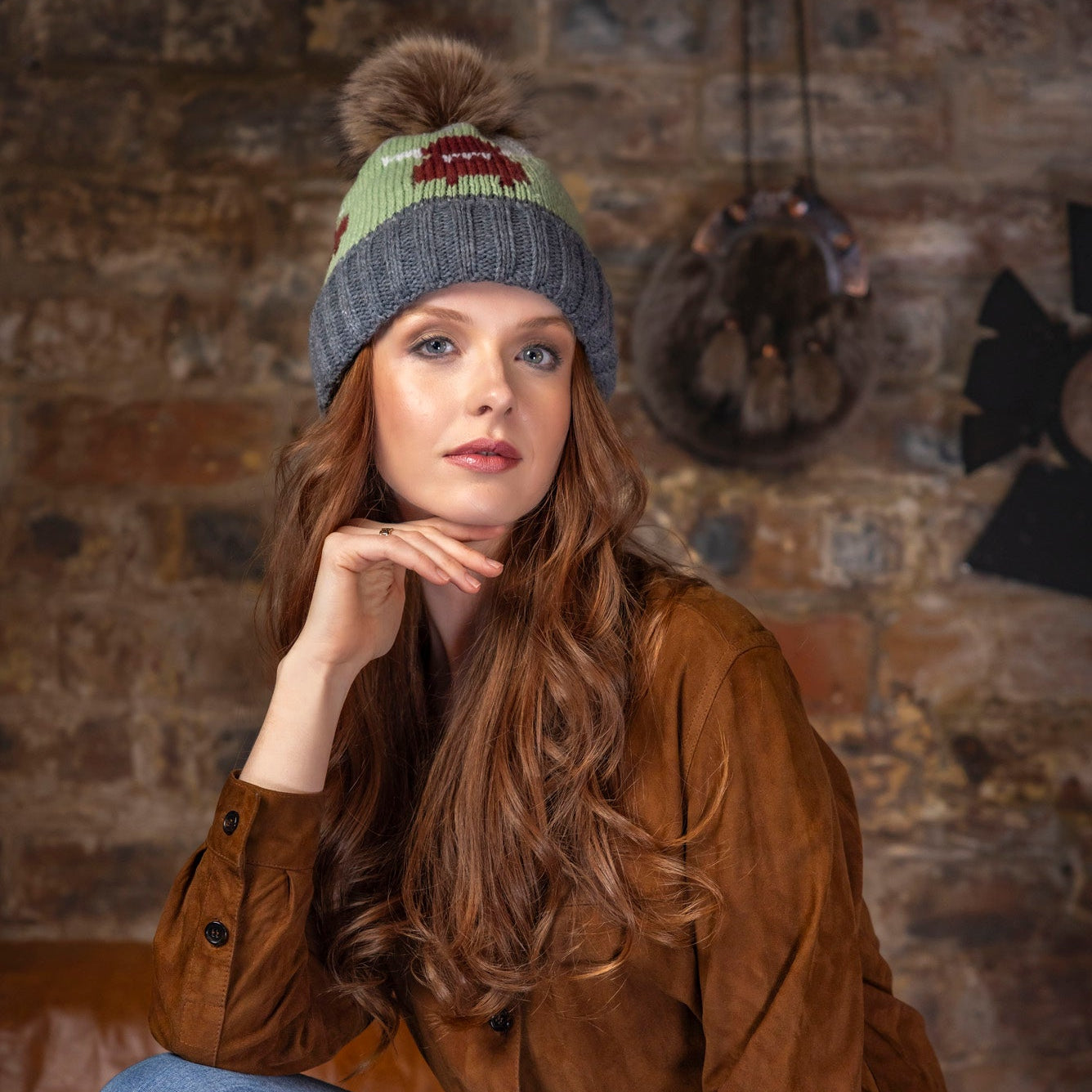 Woman wearing a brown jacket and blue jeans sitting against a brick wall.