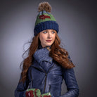 Woman wearing a blue knit hat with pom-pom, leather jacket, jeans, and colorful mittens against a gray background