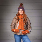 A woman posing in a brown faux-leather jacket, orange knit scarf and mittens, blue jeans, and a purple knit beanie against a gray paneled wall.