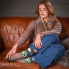 A woman sits on a brown leather couch wearing patterned socks with a green and white design, a brown knit sweater, and a plaid scarf.