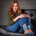 A woman with long red hair sits in a black leather lounge chair, wearing blue jeans, an olive green top, and blue crew socks with a green and white soccer pattern. The background features a gray paneled wall and a patterned rug.