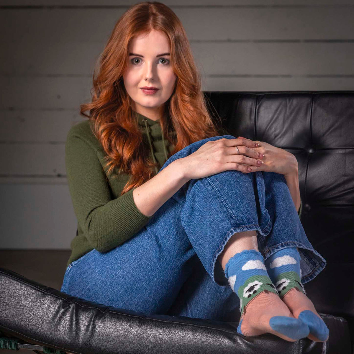 A woman with long red hair sits in a black leather lounge chair, wearing blue jeans, an olive green top, and blue crew socks with a green and white soccer pattern. The background features a gray paneled wall and a patterned rug.